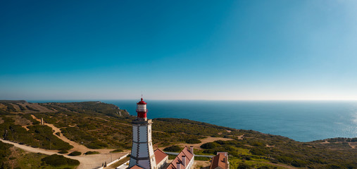 Aerial; drone view of white-red lighthouse Cabo Espichel on the edge of the earth; turquoise water of Atlantic ocean stretching to horizon; beautiful portuguese viewpoint with old historic sightseeing