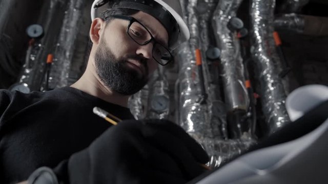 Man with beard, wearing glasses and white helmet stands in a boiler room, inspects. Against the background of hot pipes in a silver corrugation, pressure sensors. He takes notes, looks at the sensor.