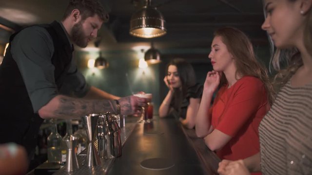 Three beautiful well dressed young girls sit by the bar and nice hipster bartender gave them one by one a cocktail