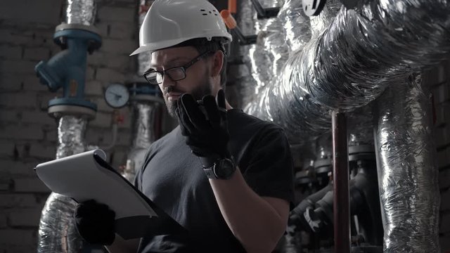 Male engineer works in boiler room and dictates audio message on smartphone. Plumber in technical room speaks in the speaker phone (speakerphone, audio message or voice recorder). Pipes on background