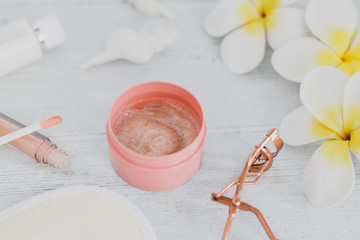 spa inspired set of scrub body lotions with make-up details such as lash curler and lipgloss surrounded by flowers and seashells on light coloured wood