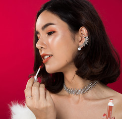 Fashion portrait of Asian Black hair tanned skin woman with strong super color red lips wearing white fur diamond earrings necklace, studio lighting red reddish background, holding liquid lipstick
