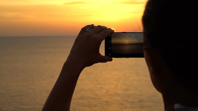 Travel concept of female hands holding a phone for taking a photo of the sunset over the sea, close up
