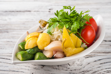 Fermented food in a plate on a white textural wooden table. Cucumbers, zucchini, garlic, marinade, tomatoes, cabbage, ramson, parsley. National Russian food pickles in brine close up and copy space