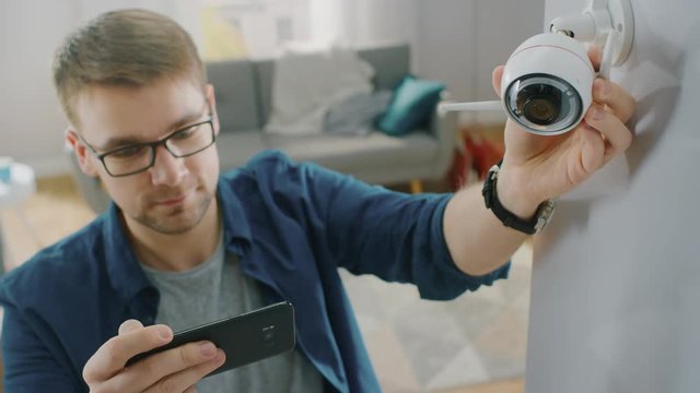 Young Man in Glasses Wearing a Blue Shirt is Adjusting a Modern Wi-Fi Surveillance Camera with Two Antennas on a White Wall at Home. He's Checking the Video Feed on his Smartphone.