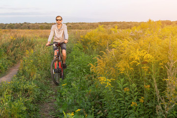 Fototapeta premium Young men on bike, cyclist in field with yellow flowers in sunlight, nature with copy space 