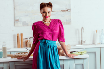 Beautiful pin up girl standing in light blue kitchen near table with different products and looking...