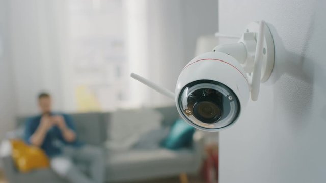 Close Up Object Shot of a Modern Wi-Fi Surveillance Camera with Two Antennas on a White Wall in a Cozy Apartment. Man is Sitting on a Sofa in the Background.