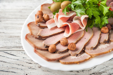 Meat and sausage sliced on a plate with nuts, garlic sauce. Pastrami, baked ham, chorizo, bacon, olives, cherry tomatoes on a plate against a white textured wooden table close-up and copy space