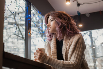 girl in a white sweater sits in a cafe and looks out the window while holding a white mug in her hands.