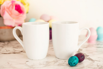 Fototapeta na wymiar Two White Mugs Mockup - Easter theme. Easter eggs. Colorful eggs in matte colors. Light marble background