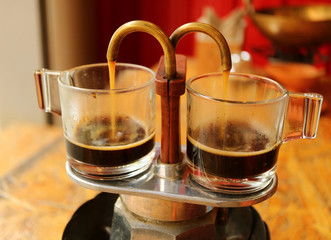 Aromatic coffee trickling out from mini retro brewing pot into a pair of glass demitasse cups 