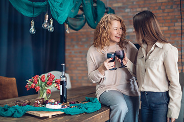 Two young women are having fun chatting around a laid table with flowers over a glass of wine at a...