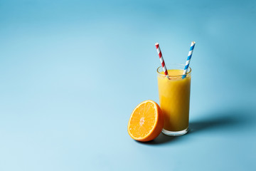 a glass of fresh juce and two paper tubes next to a lemon and an orange on a blue background with copy space