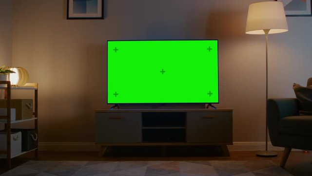 Zoom In Shot of a TV with Horizontal Green Screen Mock Up. Cozy Evening Living Room with a Chair and Lamps Turned On at Home.