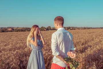 A girl with long hair in a dress standing in a wheat field with her hands covering her face. A man gives flowers behind his back. Concept of love and surprise gift, marriage proposal, engagement.