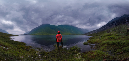 man stands by lake in red jacket in cloudy weather panorama