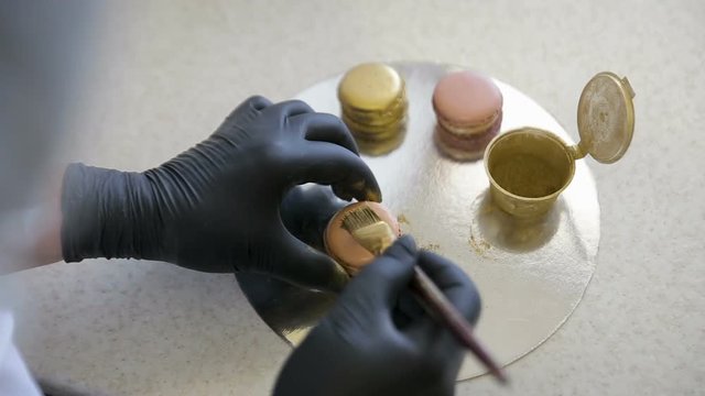 Woman hands in black gloves decorating pink macaroon with gold splash using brush in the kitchen