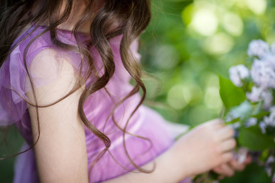 Outdoor photo of beautiful young girl in elegant dress posing in garden with blossom lilac bushes. Beautiful streaming brunet curls.  View from a back