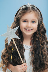 Cute little girl with positive expression, holds wand, has curly dark hair, carries wand, believes in fairy tale, isolated over blue background, wishes dreams come true. Children and magic concept