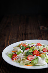 Plate of homemade fresh salad with buttermilk ranch dressing, tomatoes, broccoli, cabbage and carrots over a rustic wooden table and dark background. House Salad.