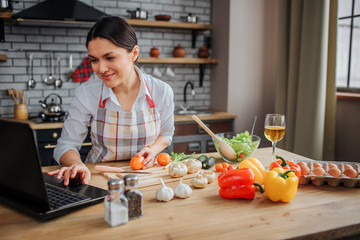 Cheerful adult woman sit at table in kitchen. She typing on keyboard and look on screen. Woman cook...