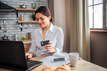 Nice cheerful woman sit at table in kitchen. She work at home. Woman hold black credit card nd type on laptop keyboard. Phone, cup and plate with croissans on table.