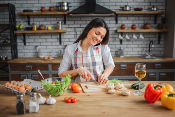 Cheerful nice woman sit at table in kitchen. She cut green onion and talk on phone. Model look down. She happy.