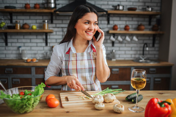 Happy gorgeous woman sit at table in kitchen. She talk on phone and smile. Woman cut green onion...