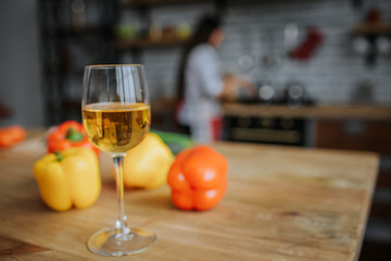 Glas of white wine stand on desk with yellow and orange peppers. Woman stand at stove in kitchen and cook.