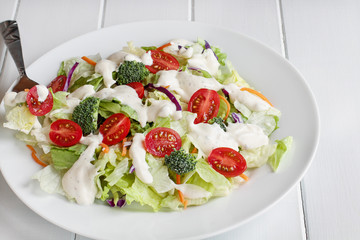 Plate of homemade fresh salad with buttermilk ranch dressing, tomatoes, broccoli, cabbage and carrots served over a white wooden table. House Salad.