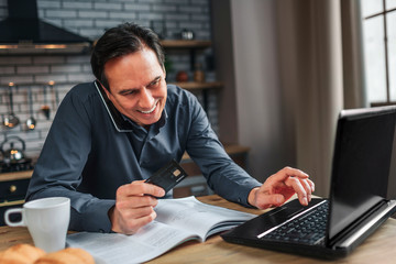 Positive man sit at table in kitchen. He talk on phone and look at papers. Man type on keyboard and smile.