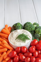 Homemade buttermilk ranch salad dressing with dill served with fresh vegetables, cherry tomatoes, baby carrots and broccoli, over a rustic white wooden table background with room for text.