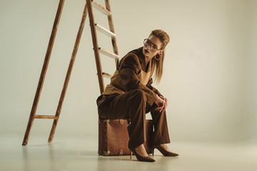 beautiful stylish woman sitting on vintage suitcase near ladder on beige