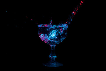 Water being poured into a glass goblet and splashing under colored lights on a black background