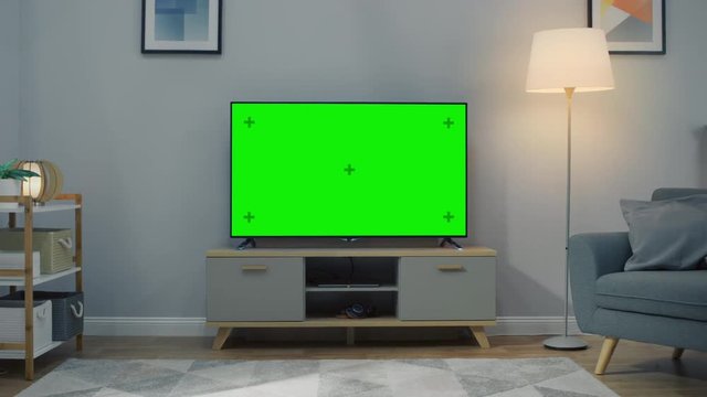 Zoom Out Shot of a TV with Horizontal Green Screen Mock Up. Cozy Living Room at Day Time with a Chair and Lamps Turned On at Home.