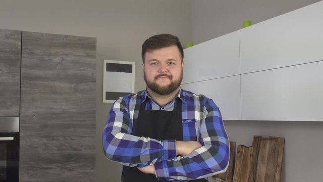 Fat bearded caucasian male cook in apron is standing in the kitchen and looking into the camera, 4K