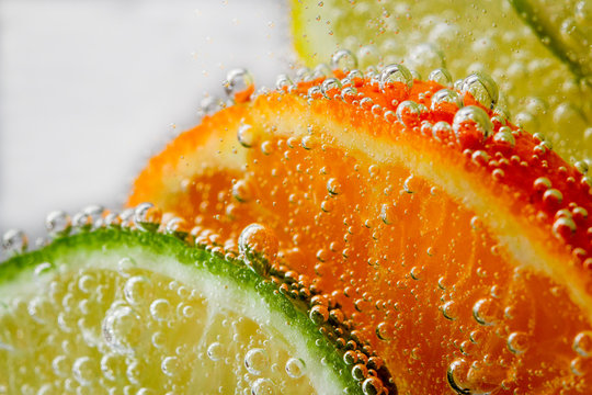 Bright, juicy orange tangerines, yellow lemons, lime green in water with air bubbles behind glass on a white background
