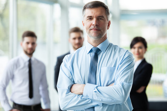 Modern mature businessman smiling and looking at camera with his colleagues in the background at office.