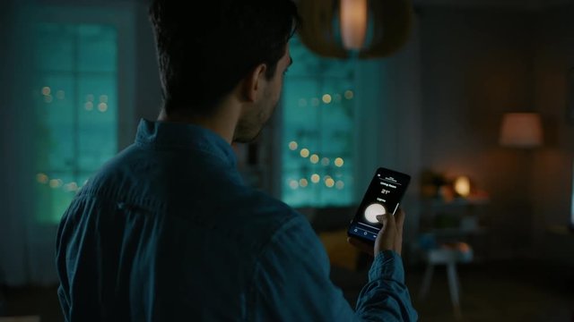 Young Handsome Man Gives a Voice Command to a Smart Home Application on His Smartphone and Lights in the Room are Being Turned On. He is Impressed by Technology. It's a Cozy Evening.