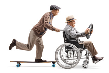 Elderly man riding a longboard and pushing a man holding a steering wheel and sitting in a...