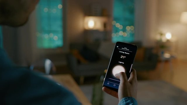 Close Up Shot of a Smartphone with Active Smart Home Application. Person is Tapping the Screen and Light is Being Turned On in the Room. It's Cozy Evening in the Apartment. 
