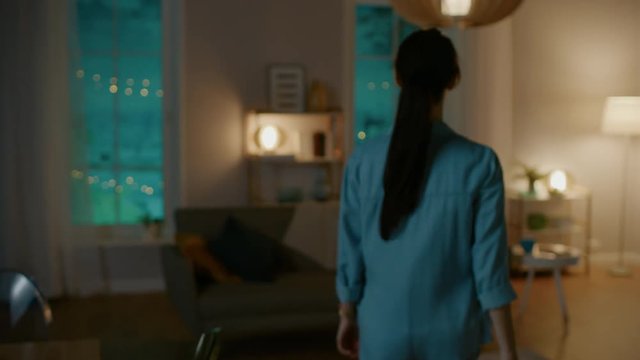 Young Beautiful Woman Gives a Voice Command to a Smart Home Application on Her Smartphone and Lights in the Room are Being Turned On. She Walks and Sits on a Couch. It's a Cozy Evening.