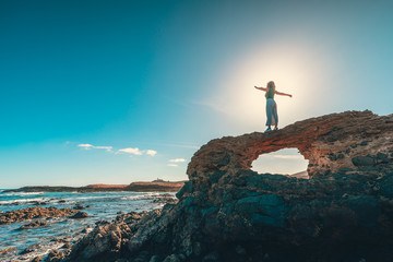 Unrecognizable female with outstretched arms standing on rough cliff near majestic sea against bright blue sky on sunny day
