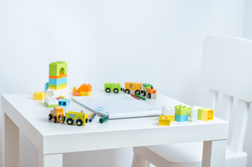White table in the bright children's room with pencils, toy train, colorful plastic block and an album with children's drawings. Children's drawing table.