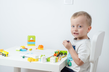 Little blond child playing with lots of colorful plastic blocks, with a toy train and draws at the table. Little boy sits on a chair at the white table.