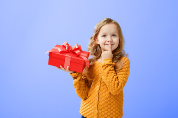Portrait little girl on a blue background. The child looks dreamily up and holds a red box. The...