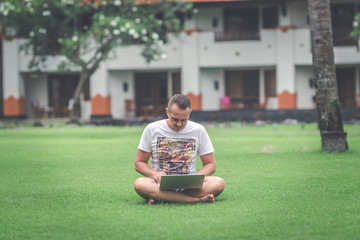 Fototapeta premium Young man in the green tropical park working online with laptop. Freelancer man on Bali island concept.