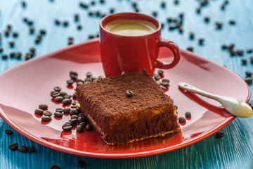 Tiramisu cake on a red plate, near a cup of coffee, photographed on a light wooden background.