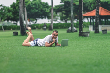 Fototapeta premium Young man in the green tropical park working online with laptop. Freelancer man on Bali island concept.